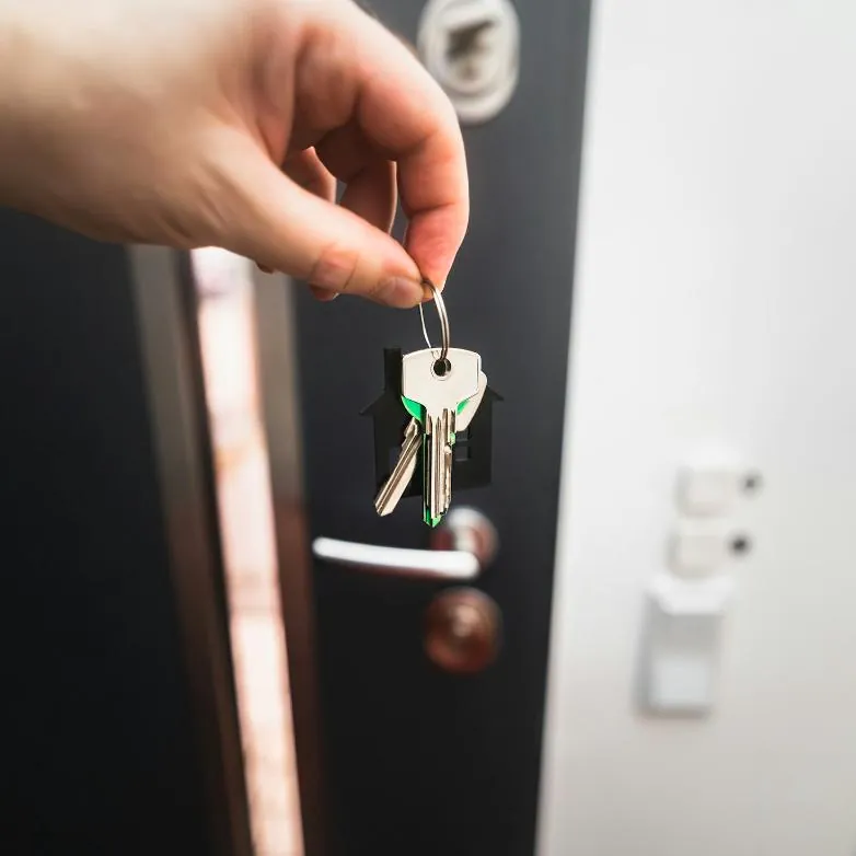 A hand holding a set of house keys on a house-shaped key ring beside a front door